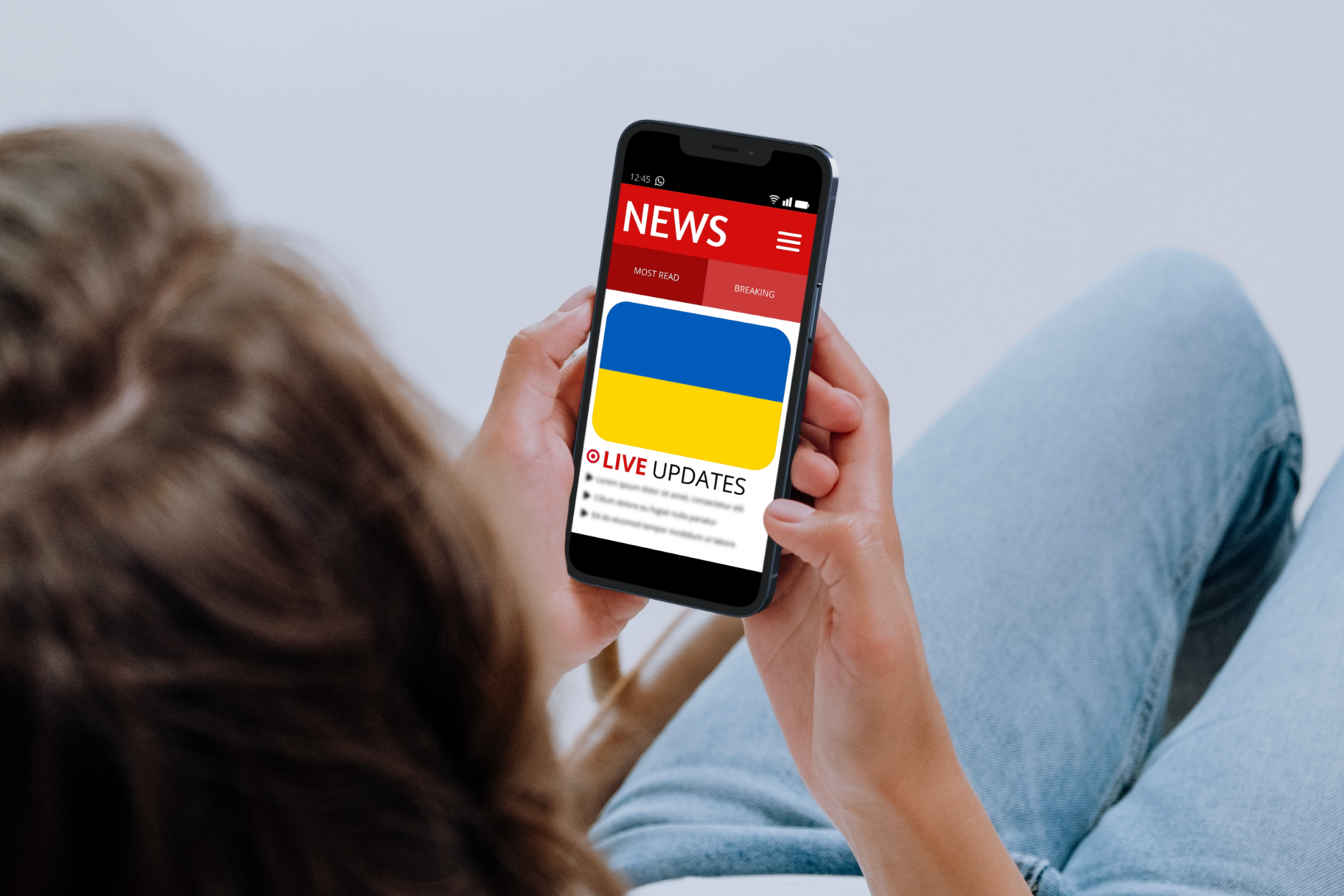 Image shows a young woman (18-30) holding a mobile phone. On the phone screen is a news app - showing live updates of the Russian Invasion of Ukraine. Overconsumption of news during crises can be negative to our wellbeing.
