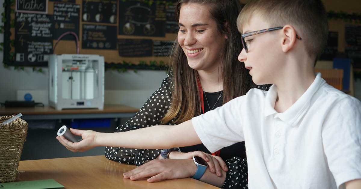 A teacher with a primary school pupil looking at a 3D printed object with a 3D printer in the background of the classroom.