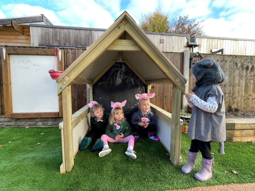 three children dressed as little pigs sit inside the essentials play house as another child dressed as a wolf pretends to howl outside